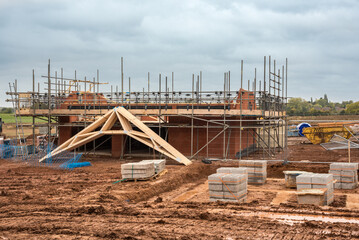 Fototapeta premium Family house construction site. Building red brick house surrounded by scaffolding with wooden roof truss. Energy-efficient homes estate development. Works on new residential properties for homebuyers