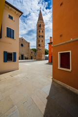 Obraz premium A narrow European alleyway with colorful buildings and a tall stone tower under a blue sky. Caorle, Metropolitan City of Venice, Veneto, Italy