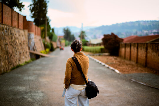 Woman with a shoulder bag walking down an empty street lined with walls and trees. Kigali, Rwanda