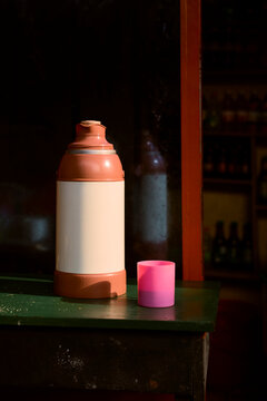 A large brown thermos and pink cup on a dark green table in a dimly lit room. Kigali, Rwanda