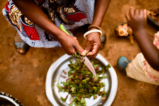 Hands of a person cutting leafy greens over a bowl on a dirt ground, child nearby. Kigali, Rwanda