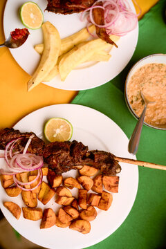 Two plates of grilled skewers and plantains with a side of sauce on a colorful tablecloth. Kigali, Rwanda