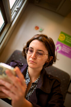 Individual in glasses absorbed in phone on a train, seated by window with colorful signage. Belgium