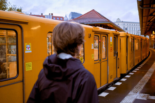 Person in a black jacket waits for a yellow subway train at an outdoor urban platform. Germany