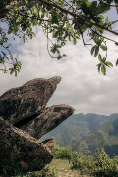 Large jagged boulders with a scenic mountain view framed by tree branches and cloudy sky. Mountains of Ta Xua, Bac Yen District, Son La, Vietnam