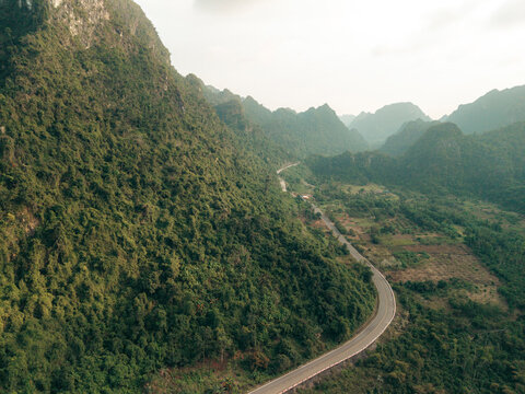 A winding road cuts through lush green mountains under a cloudy sky, creating a scenic vista. Lan Ha Bay, Cat Ba, Vietnam