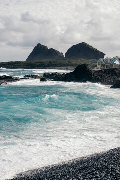 Blue ocean waves crash against rocky dark coast under cloudy skies and distant hills. Taiwan, Taitung County, Chenggong Township