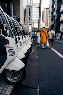Row of parked scooters on urban street beside people walking in bright coats Tokyo, Japan