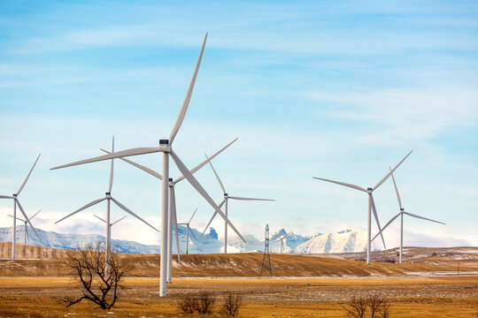 Wind turbines stand tall against a snowy mountain backdrop under a clear blue sky. Calgary, Canada