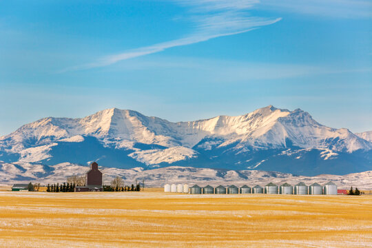Snow-capped mountains with a field and silos under a clear blue sky. Calgary, Canada