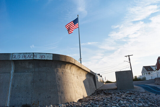 An American flag flies atop a concrete wall with a road and houses in the background. Marshfield,, MA, USA