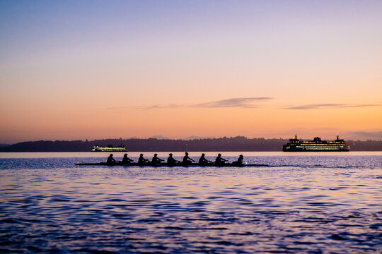 Rowers glide across the calm water at dusk with ferries on the horizon under a colorful sky. WA, USA