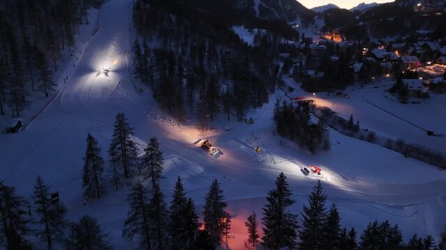 Aerial view of snow-covered ski slopes with snow groomers and town lights creating a warm glow against the cold, dark winter evening, Claviere, Piedmont, Italy.