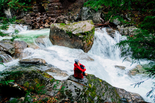 Person in red sits by a rushing river surrounded by lush green forest and large rocks. Deception Falls, Pacific Northwest, WA, USA