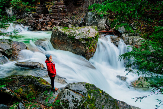 A person in a red jacket stands by a cascading waterfall in a lush forest setting. Deception Falls, Pacific Northwest, WA, USA
