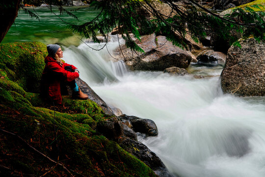 Person in red jacket sits by a lush, mossy riverbank, admiring the rushing water. Deception Falls, Pacific Northwest, WA, USA