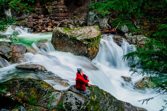 A person in red admires a flowing waterfall surrounded by rocks and lush greenery. Deception Falls, Pacific Northwest, WA, USA