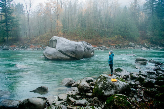 Person in a blue jacket standing by a misty river bank with rocks and trees in the background.  Deception Falls, Pacific Northwest, WA, USA