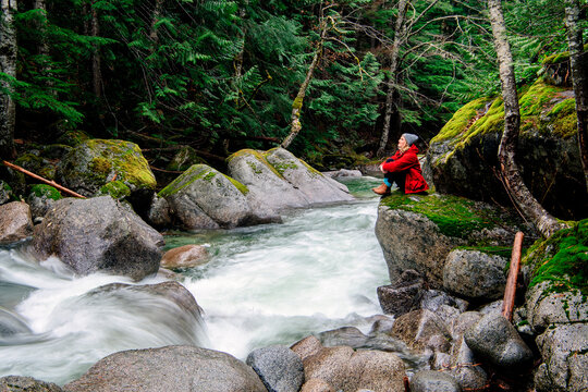 Person in red jacket sitting by a rushing river in a lush green forest. Deception Falls, Pacific Northwest, WA, USA