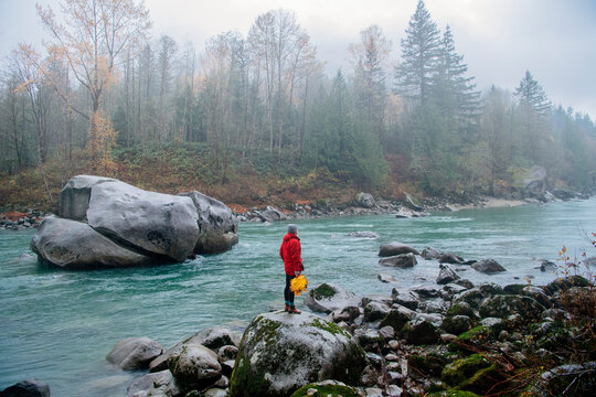 A person in a red jacket stands by a flowing river surrounded by rocks and trees in autumn. Deception Falls, Pacific Northwest, WA, USA