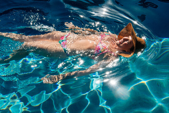 Woman in floral bikini and sunhat floats joyfully on bright blue pool water. USA