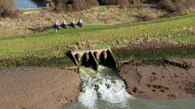 Aerial view of water gushing from the Hobhole Pumping Station outlet, contrasting against the muddy banks and verdant greenery, Boston, United Kingdom.