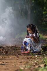 A small Asian girl is sitting while reading or writing a book outside the house, in a typical rural atmosphere.