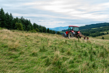 A red farm tractor with an attachment mowing thistles on a grassy hillside overlooking green pastures and distant mountains under an overcast sky