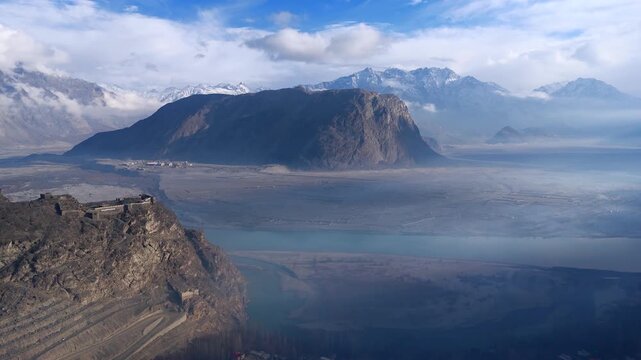 Aerial view of the historic Altit Fort perched on a cliffside, overlooking a serene river valley surrounded by imposing mountains, Skardu, Gilgit Baltistan, Pakistan.