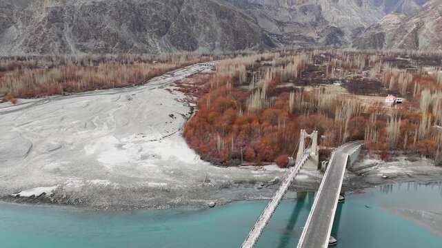 Aerial view of a bridge over a wide, stony riverbed, surrounded by autumn-colored trees against a backdrop of rugged mountains, Daghoni Balgar, Pakistan.
