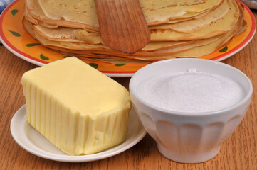Morceau de beurre et ramequin de sucre en poudre devant une assiette de cr&ecirc;pes bretonnes avec une spatule en bois