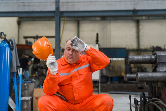 Exhausted industrial worker taking break wiping sweat from forehead after working hard in hot factory. Tired technician holding safety helmet feeling heat stroke during maintenance in workshop