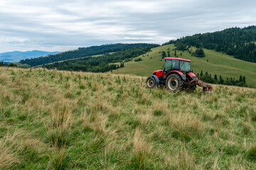 A red tractor with a harrow on a grassy hillside, amidst forested mountains and green valleys under an overcast sky, mowing thistles