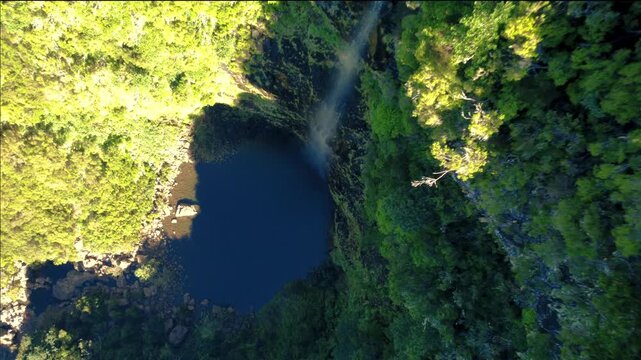 Aerial top down view of Lagoa do Vento waterfall in Madeira, drone descending over hidden lagoon in deep shadows with bright sunlight hitting mountain cliffs, dramatic high contrast landscape.
