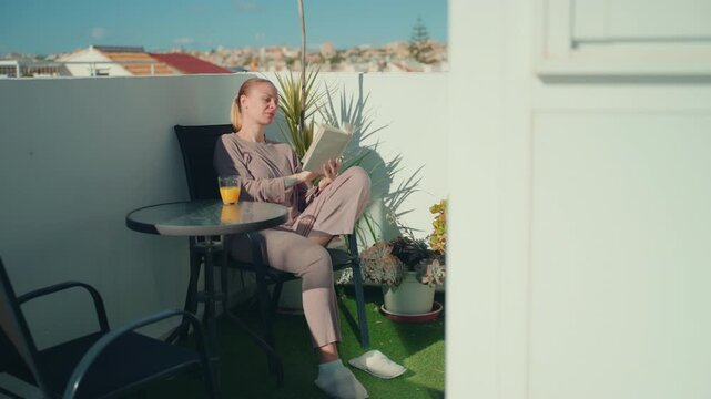 Focused woman read book and drink fresh orange juice on sunny balcony on terrace in daylight. Education, learning, self development and knowledge concept. Calm outdoor study environment.