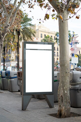 Blank vertical advertising billboard standing prominently on a city sidewalk next to a large tree and several trash cans, offering a clear space for mockup designs