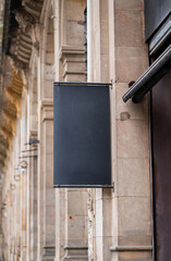 Obraz premium Blank black shop sign mockup hanging on historic stone wall, providing space for branding, advertising, and street navigation in urban area