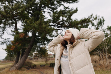 Outdoor woman wearing a beige puffer coat and knit beanie, leaning back with arms raised, enjoying fresh air among trees in a calm park setting.
