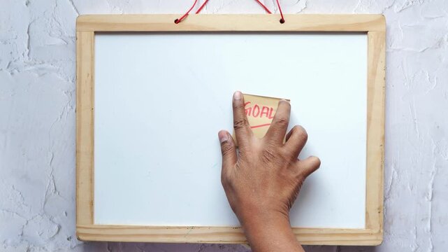 Close-up slow-motion video of hand sticking 'Goal' note on whiteboard, emphasizing planning and achievement concept
