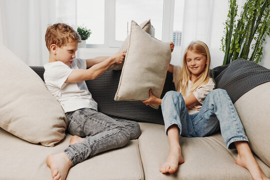 Two children having fun on a couch with pillows, playfully fighting during a cozy moment in a bright living room filled with natural light and plants.