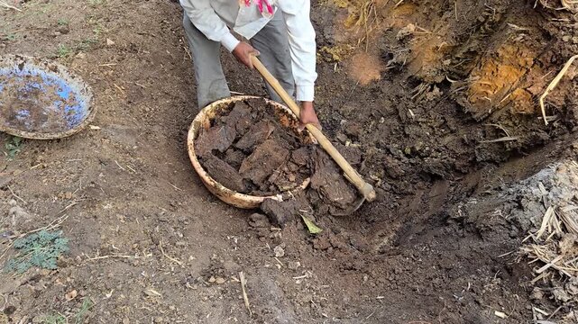 A worker uses a wooden tool to dig dark soil into a large circular tub. They are positioned in a dirt pit or field, working manually among piles of earth and debris in an outdoor setting.