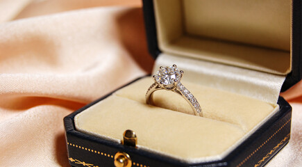 Close-up of diamond engagement ring in elegant box with round-cut stone and smaller diamonds on band. Luxurious romantic scene with satin fabric background, symbolizing love and celebration