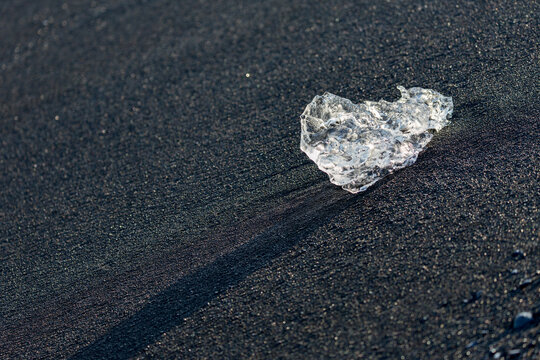 A crystal‑clear shard of ice glows against the dark volcanic sand of Diamond Beach near J&ouml;kuls&aacute;rl&oacute;n glacier lake as soft sunrise light stretches across Southern Iceland&rsquo;s rugged shoreline