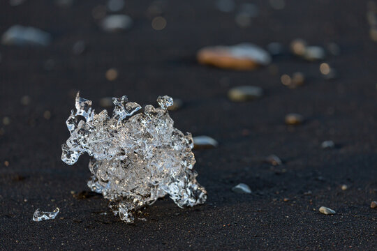 A crystal‑clear shard of ice glows against the dark volcanic sand of Diamond Beach near J&ouml;kuls&aacute;rl&oacute;n glacier lake as soft sunrise light stretches across Southern Iceland&rsquo;s rugged shoreline