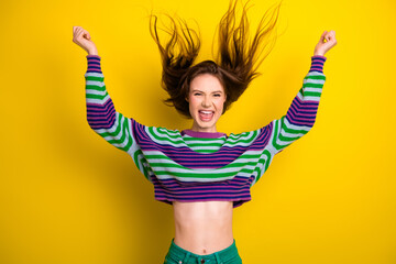 Young woman jumps with arms raised wearing striped sweater against yellow background expressing...