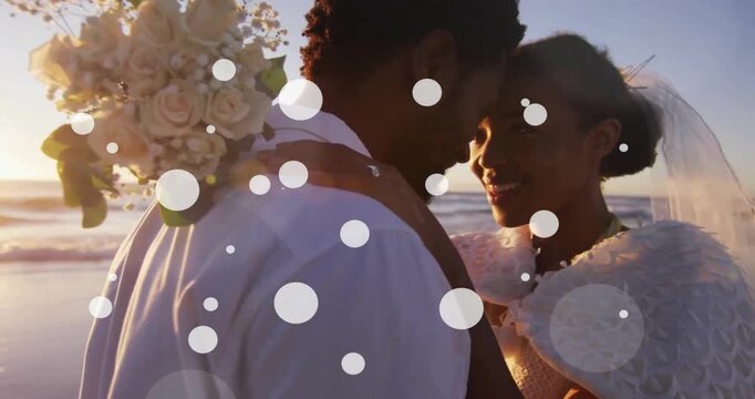 Animation of white spots over happy african american bride and groom embracing on beach at wedding