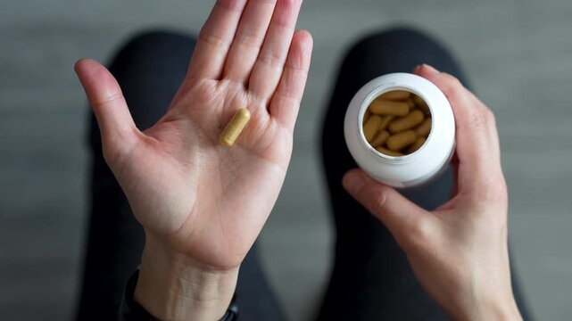 Top View of Woman Pouring Supplement Capsules Into Palm. A woman pours yellow medicine capsules from a white plastic bottle into her hand. Healthy habits, pharmaceutical treatment, and vitamin intake