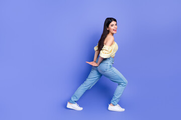 Young woman in yellow off shoulder blouse and jeans smiles while posing against a purple background