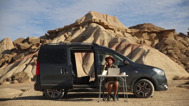 A young man wearing a hat works on his laptop and smartphone at a camping table next to his camper van in the middle of the Bardenas Reales desert, Spain.