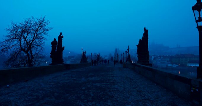 Quiet winter morning on Charles Bridge in Prague with only a few people crossing. Cold blue atmosphere and soft dawn light create a peaceful and cinematic city scene.